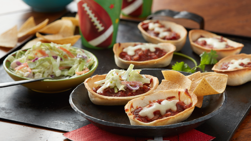 Mini bols de tortillas tendres Old El Paso remplis de porc barbecue et de fromage fondus en forme de bande de football sur une planche de service en bois noir.