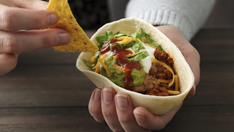 A hand holding an Old El Paso Soft Tortilla Bowl with guacamole, ground beef, sour cream, cheese, lettuce and refried beans